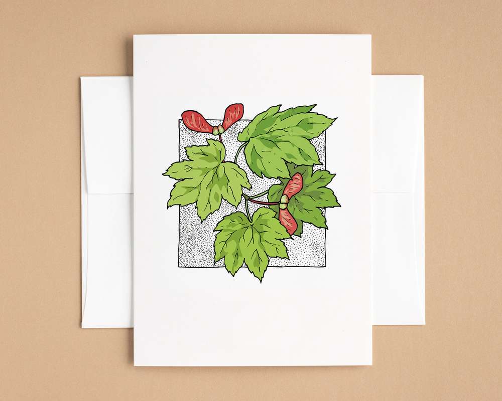 A vertical white card depicts 4 maple leaves and two red maple seeds. The card sits on top of a white envelope, which lies on top of a brown backdrop.